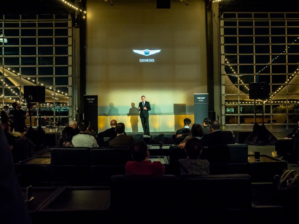 A speaker on a lit stage under a Genesis logo, facing an audience seated in lounge furniture