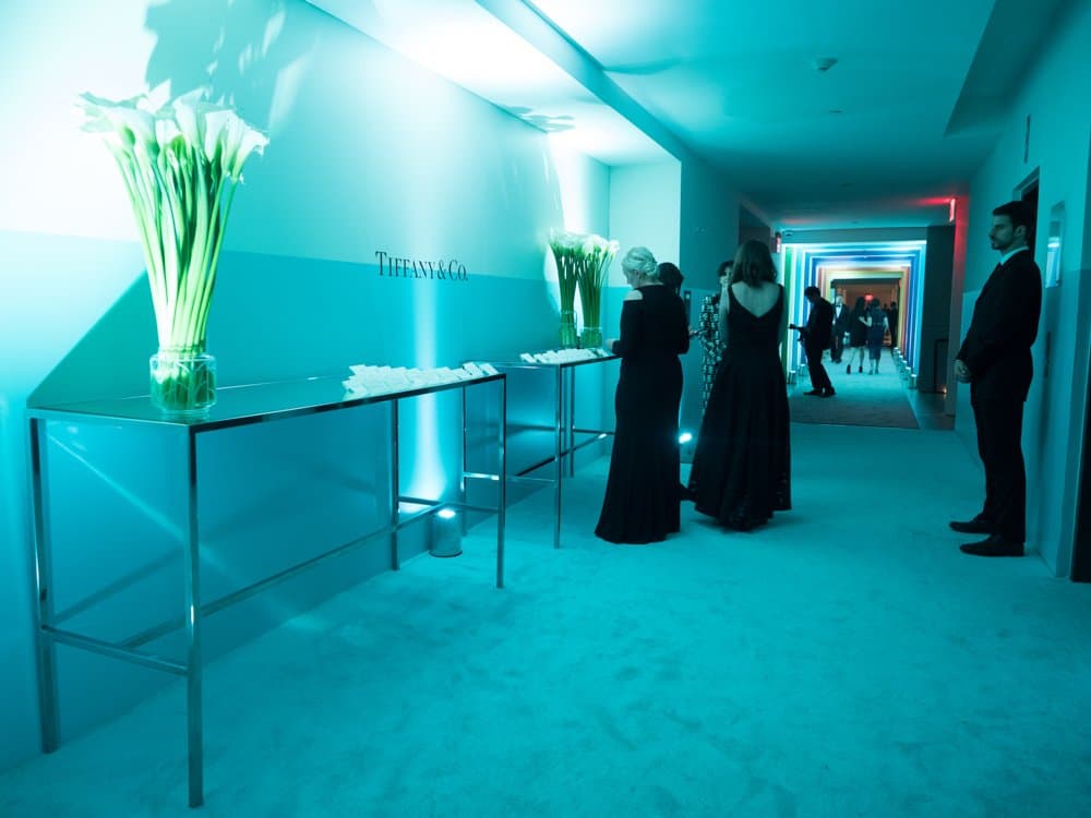 Guests in black tie along a Tiffany-blue corridor lined with escort card tables