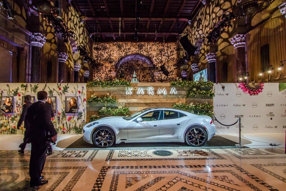 A white Karma car on a plinth beside a step-and-repeat in a marble hall