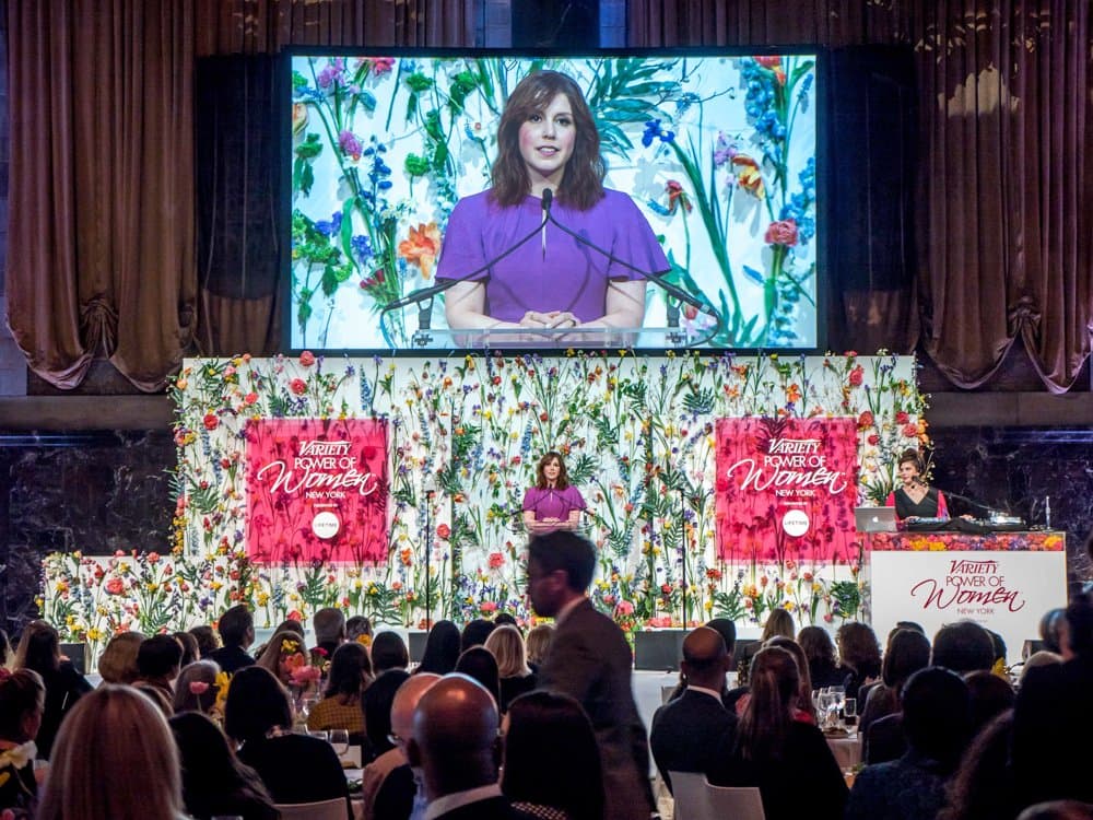 A speaker at a podium in front of a floral backdrop and a large screen, facing a seated audience